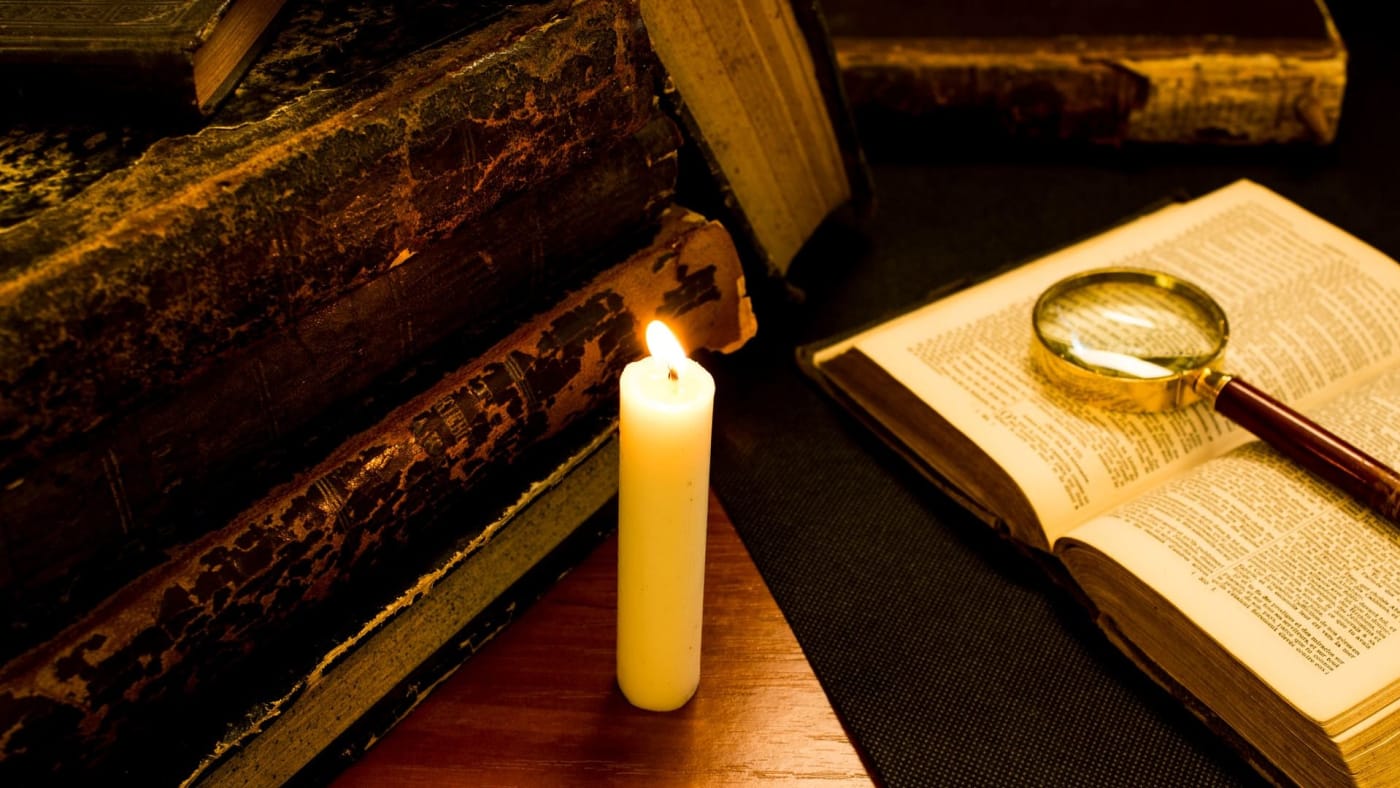 Pilha de livros antigos ao lado de uma vela acesa e um livro aberto com uma lupa sobre as páginas.