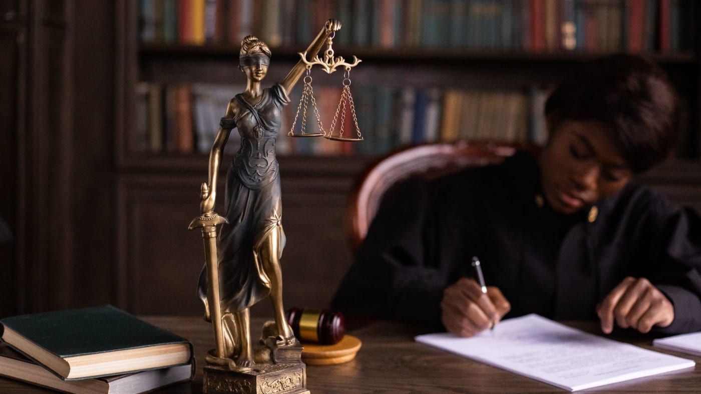 Estátua da Justiça sobre mesa de madeira com livros e uma mulher escrevendo ao fundo.
