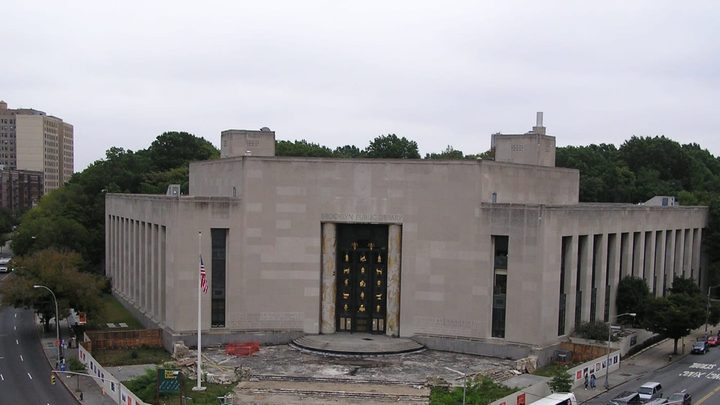 Vista aérea da fachada monumental da Biblioteca Pública do Brooklyn com árvores ao fundo e rua em frente.