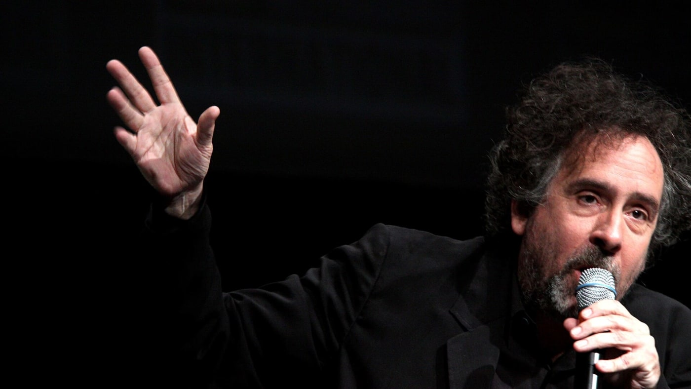 Tim Burton segura um microfone e gesticula com a mão durante uma apresentação.