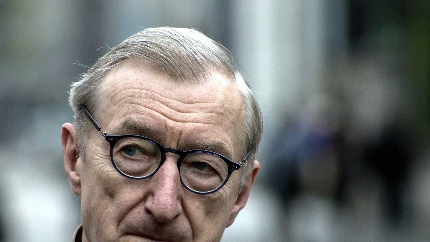 Julian Barnes de óculos e casaco bege olhando em direção à câmera.