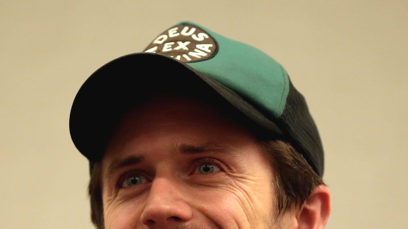 Pierce Brown sorri usando boné e camisa escura diante de um microfone em um evento.