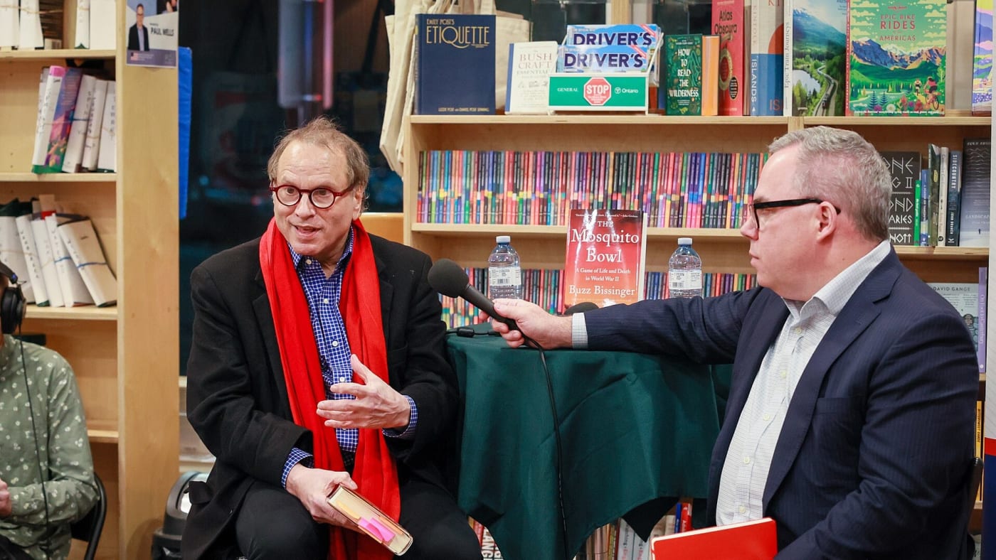 Buzz Bissinger com cachecol vermelho fala ao microfone durante evento de lançamento de seu livro.