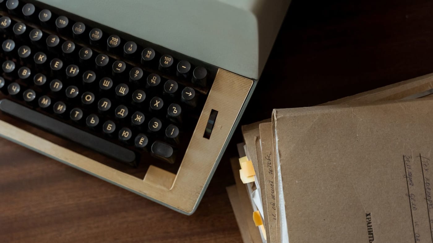 Máquina de escrever vintage ao lado de pastas com papéis sobre uma mesa de madeira.