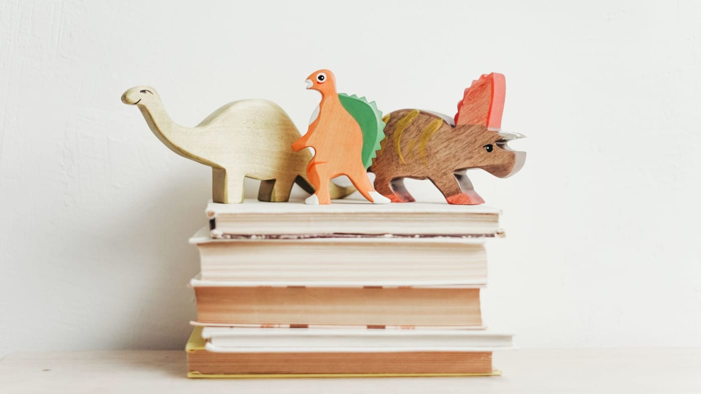 Pilha de livros com brinquedos de madeira em formato de dinossauros sobre eles contra uma parede branca.