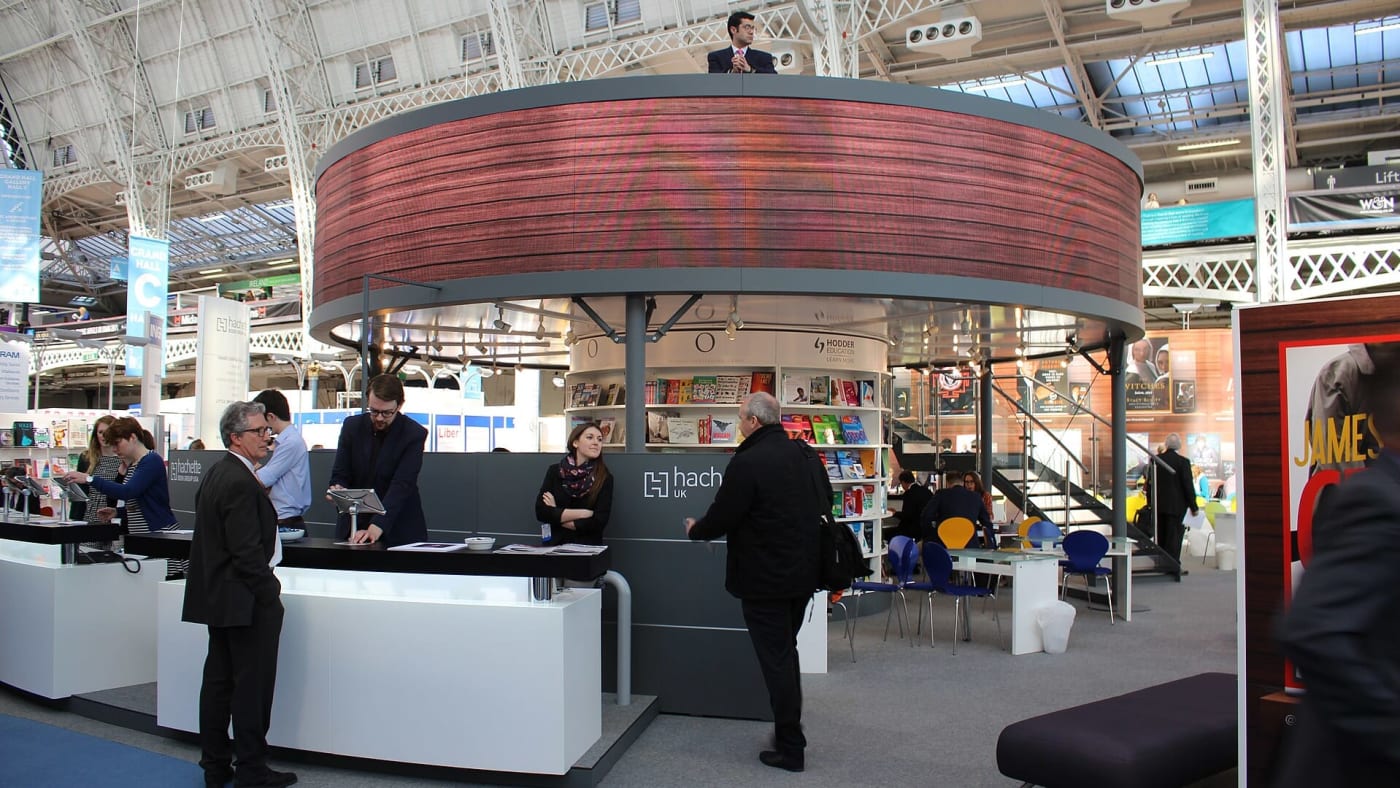Estande circular da editora Hachette com livros expostos e funcionários em feira literária.