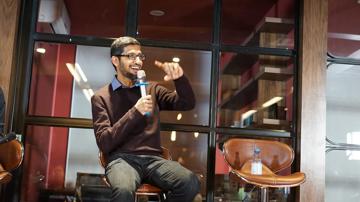 Sundar Pichai sorri enquanto fala ao microfone e gesticula sentado em evento