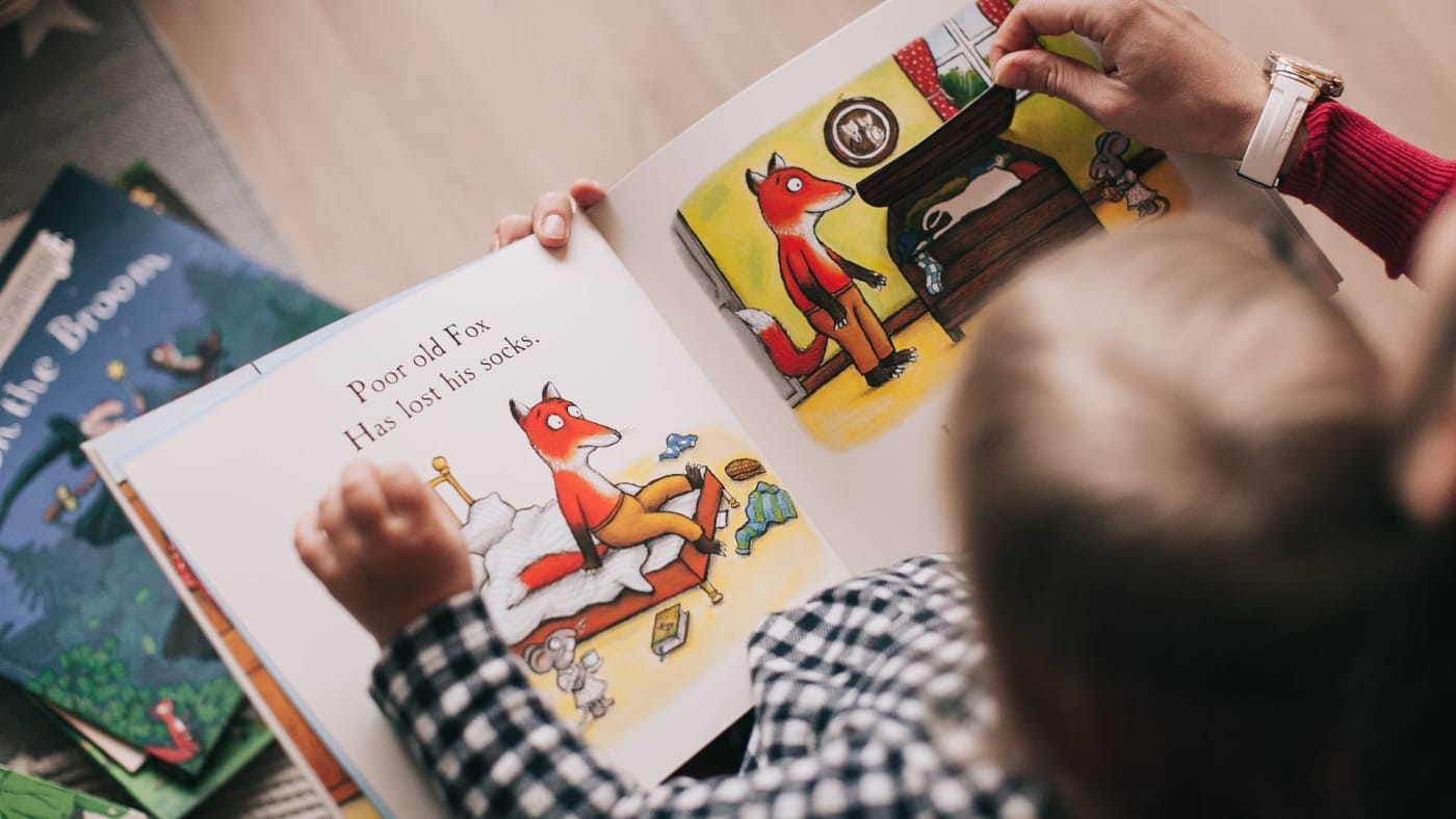 Criança e adulto segurando e lendo um livro infantil ilustrado aberto.
