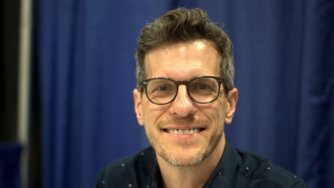 Brian Selznick sorri usando óculos de armação escura e camisa azul com pequenas estrelas brancas.