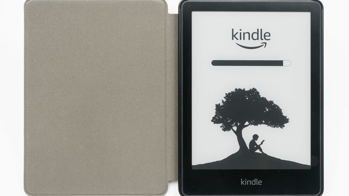 Leitor digital Kindle com capa cinza aberta exibindo a tela inicial sobre fundo branco.