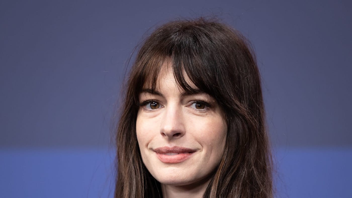 A atriz Anne Hathaway sorri em frente a um painel azul em evento.