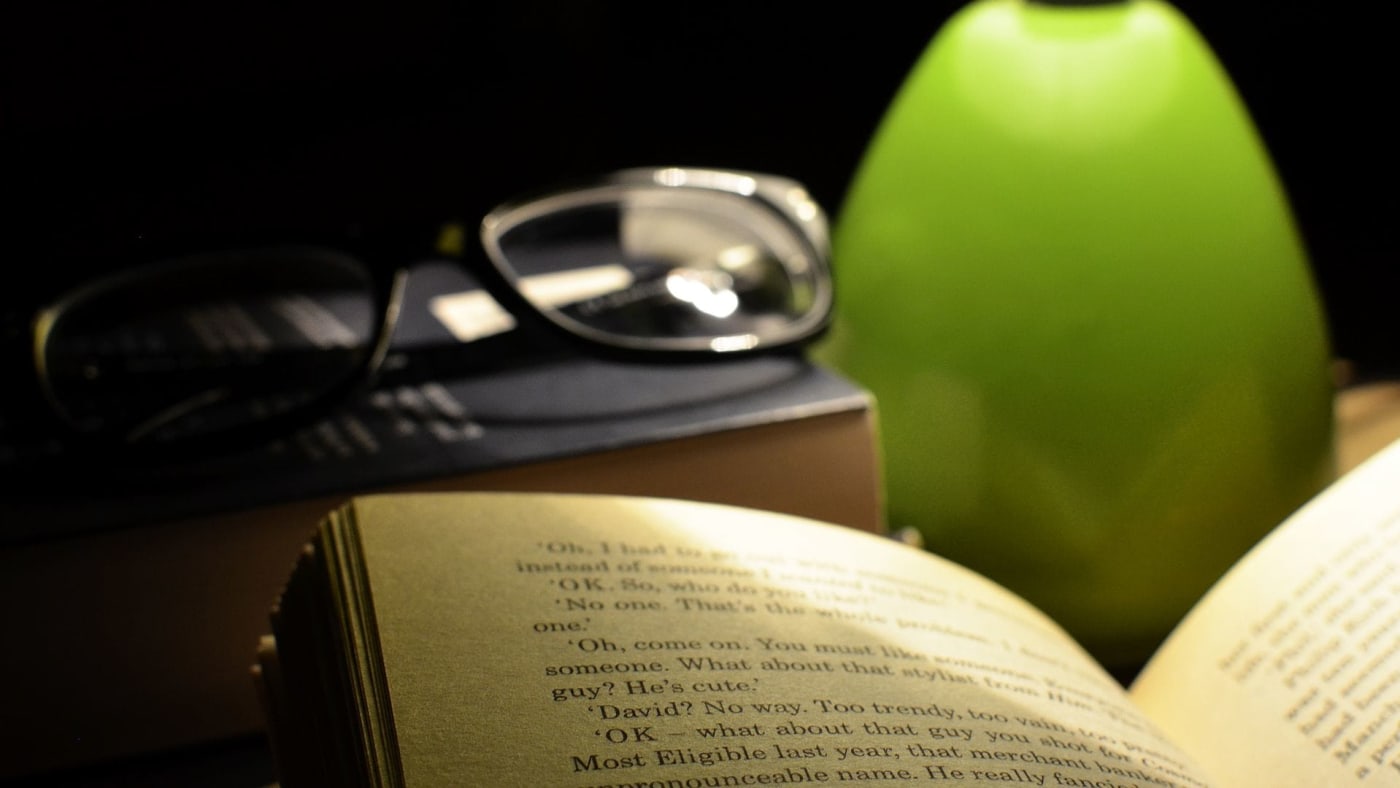 Livro aberto iluminado por uma luminária verde, com óculos apoiados sobre outro livro ao lado.
