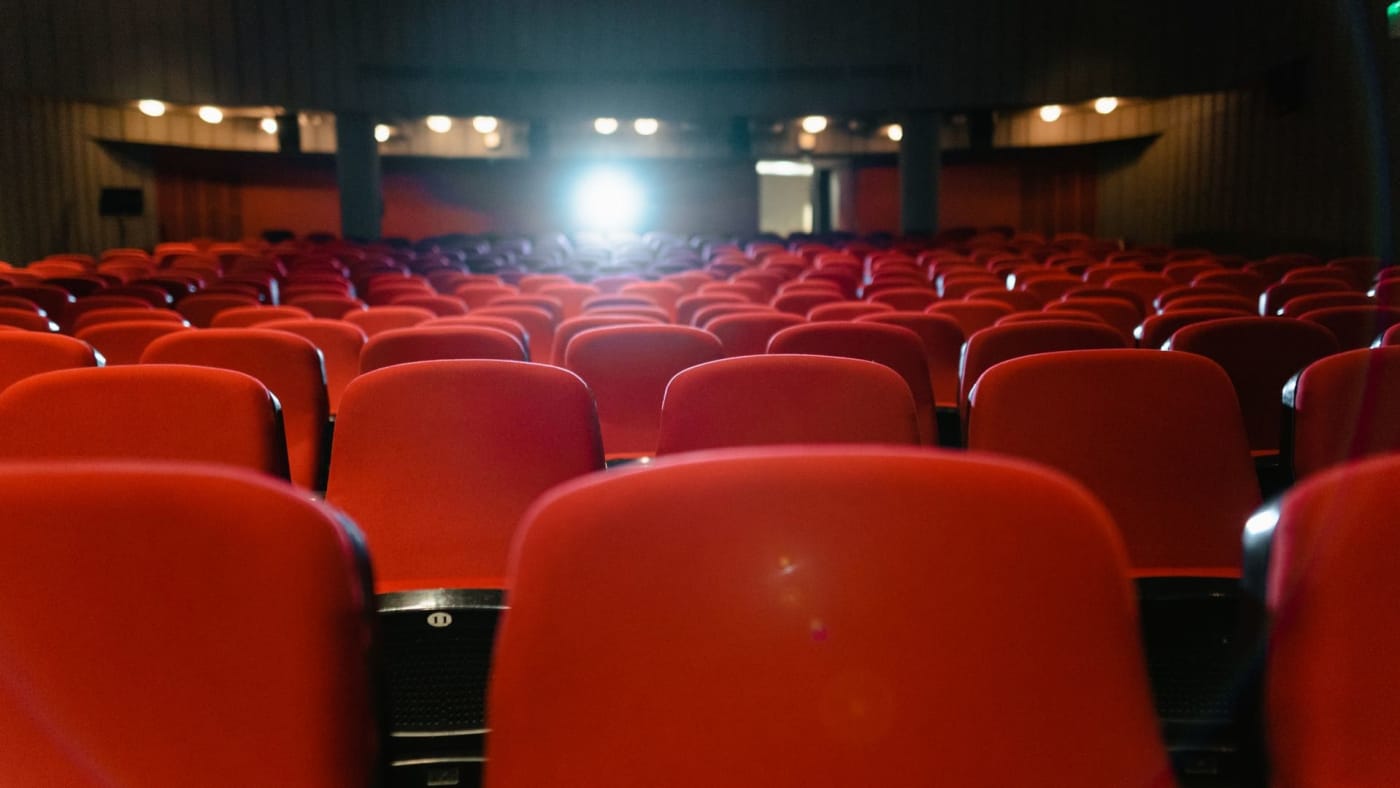 Fileiras de poltronas vermelhas em uma sala de cinema escura e vazia.