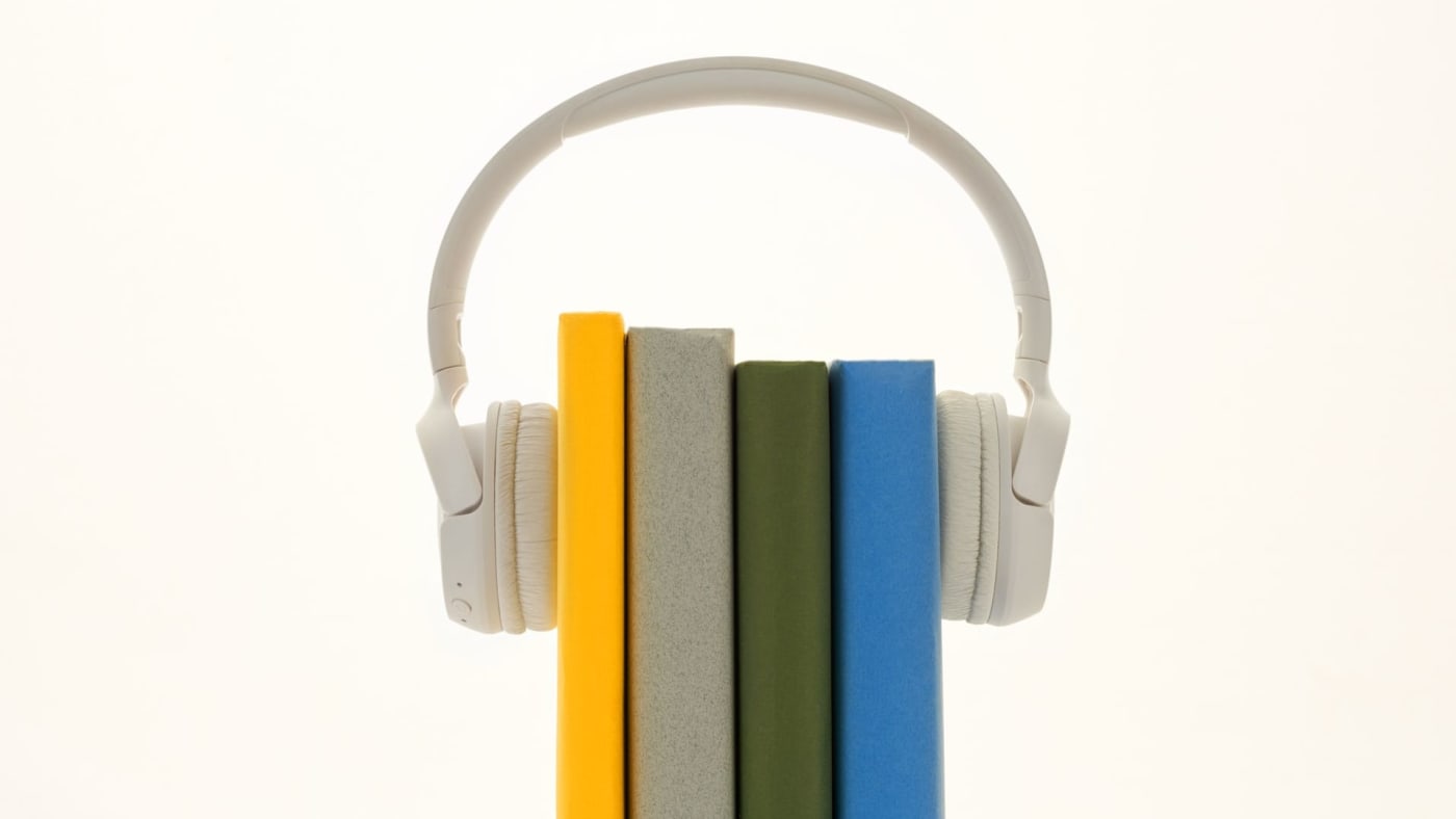 Fones de ouvido brancos encaixados sobre quatro livros coloridos em pé contra um fundo branco.