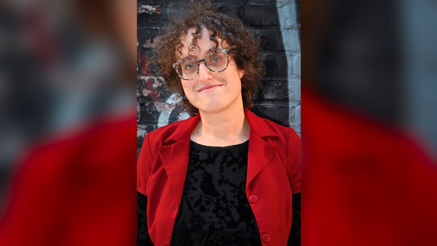 Jane Schoenbrun, de óculos e blazer vermelho, diante de um muro grafitado