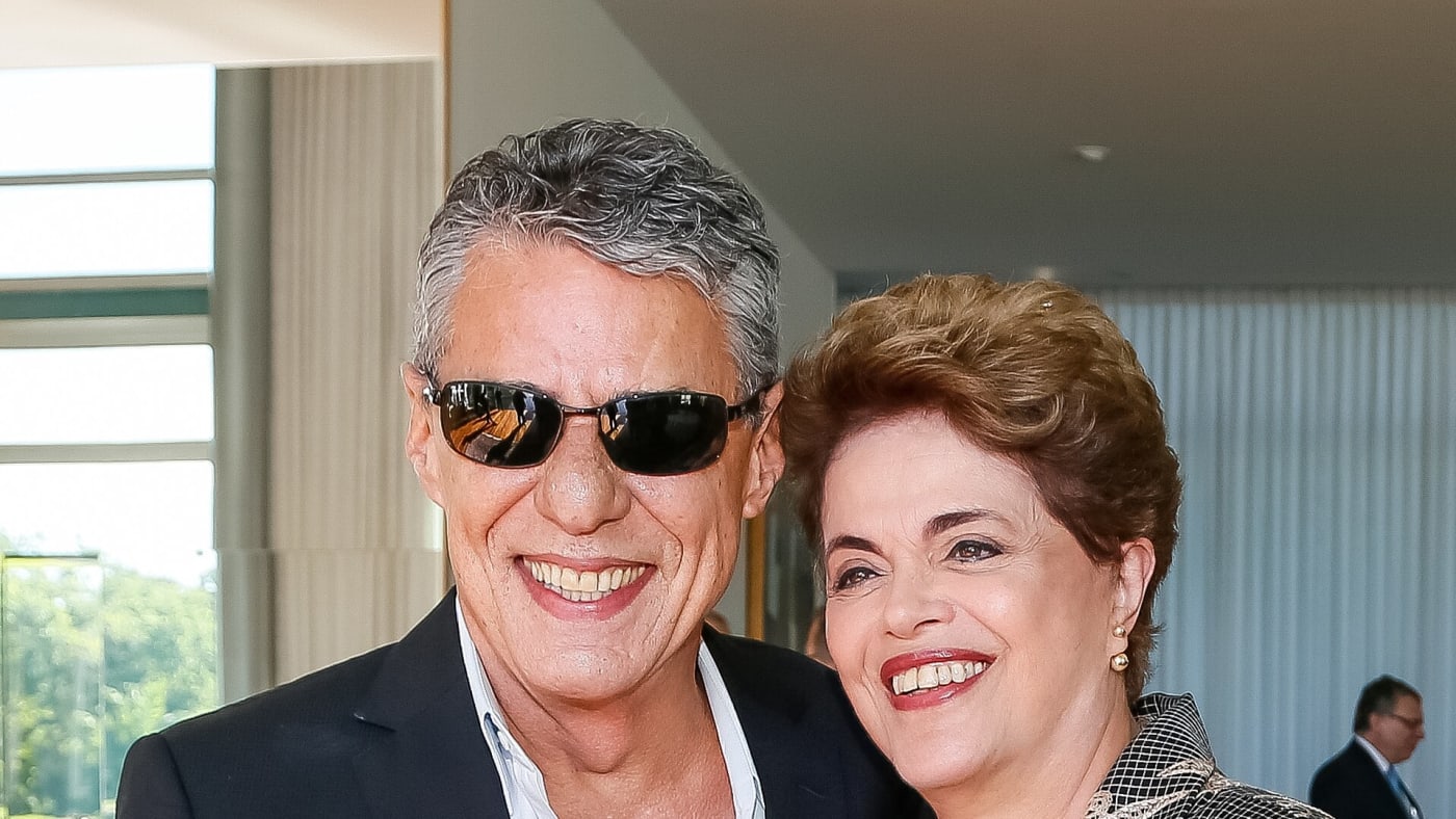 Chico Buarque de óculos escuros sorri ao lado de Dilma Rousseff