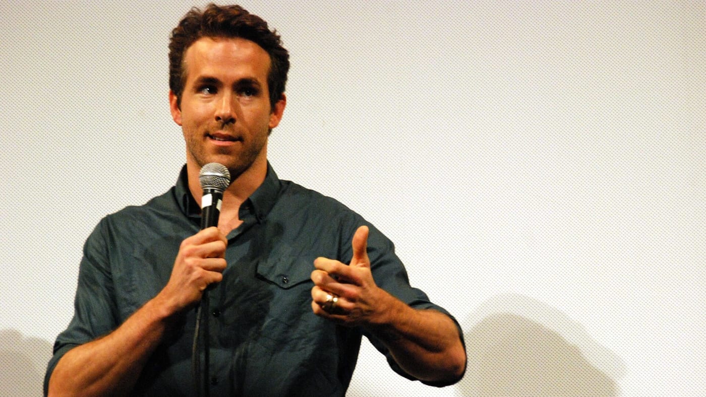 Ryan Reynolds segura um microfone e faz sinal de positivo durante evento
