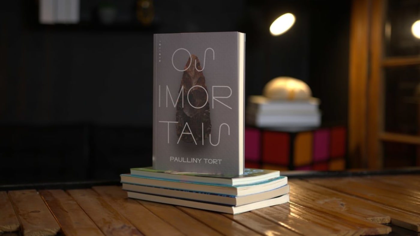 Exemplar do livro 'Os Imortais', de Paulliny Tort, apoiado sobre uma pilha de livros em cima de uma mesa de madeira.