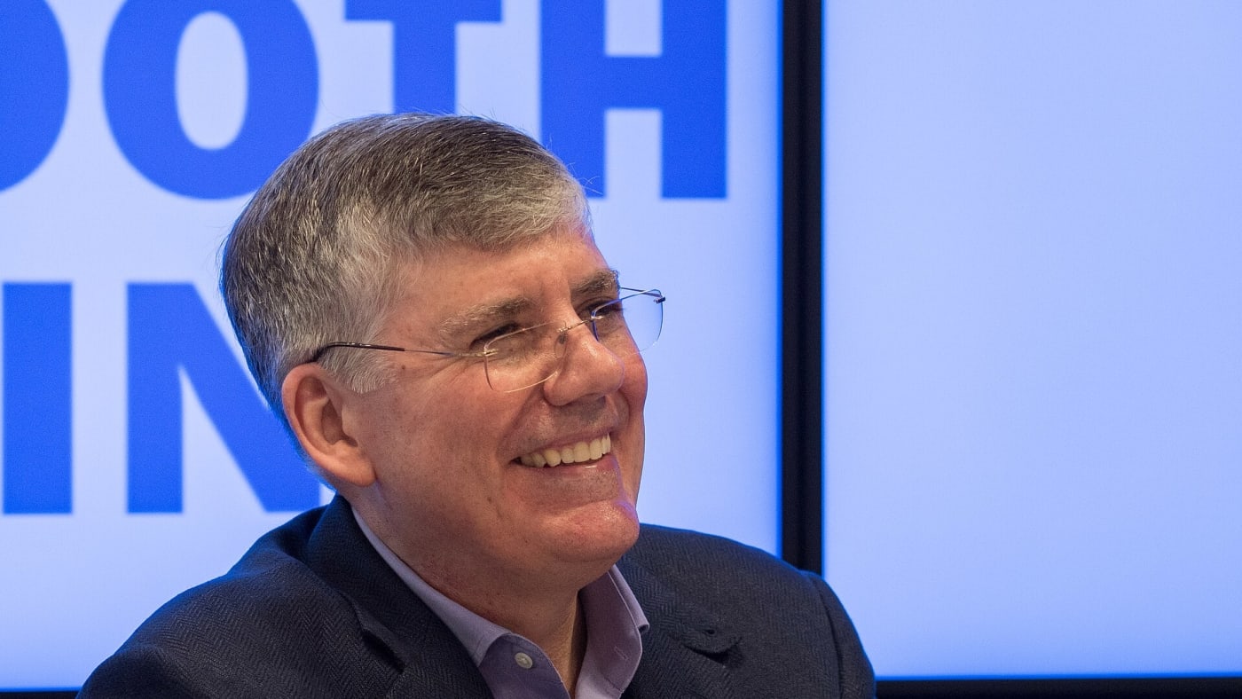 O escritor Rick Riordan sorri durante um evento, usando óculos e paletó azul.