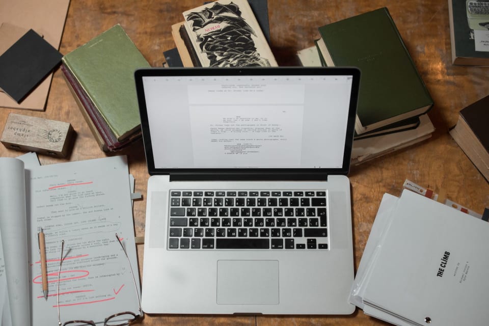 Mesa de trabalho com páginas de livro impressas, lápis e provas espalhadas sob luz lateral