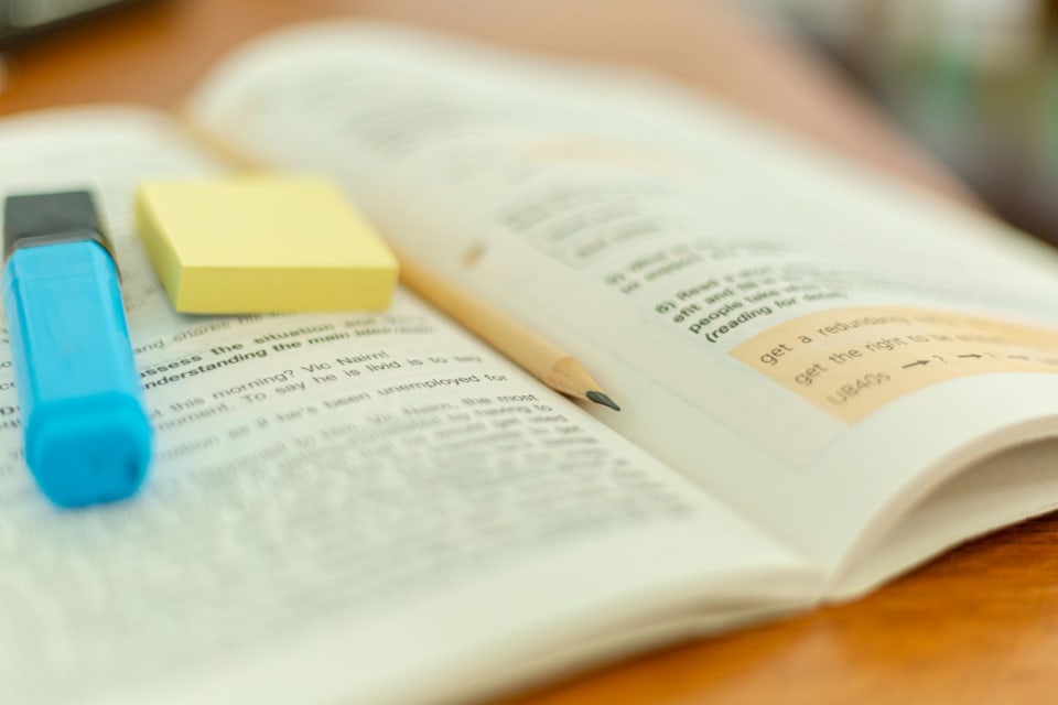 Livro aberto sobre uma mesa de madeira, com marca-texto, bloco adesivo e lápis sobre as páginas.