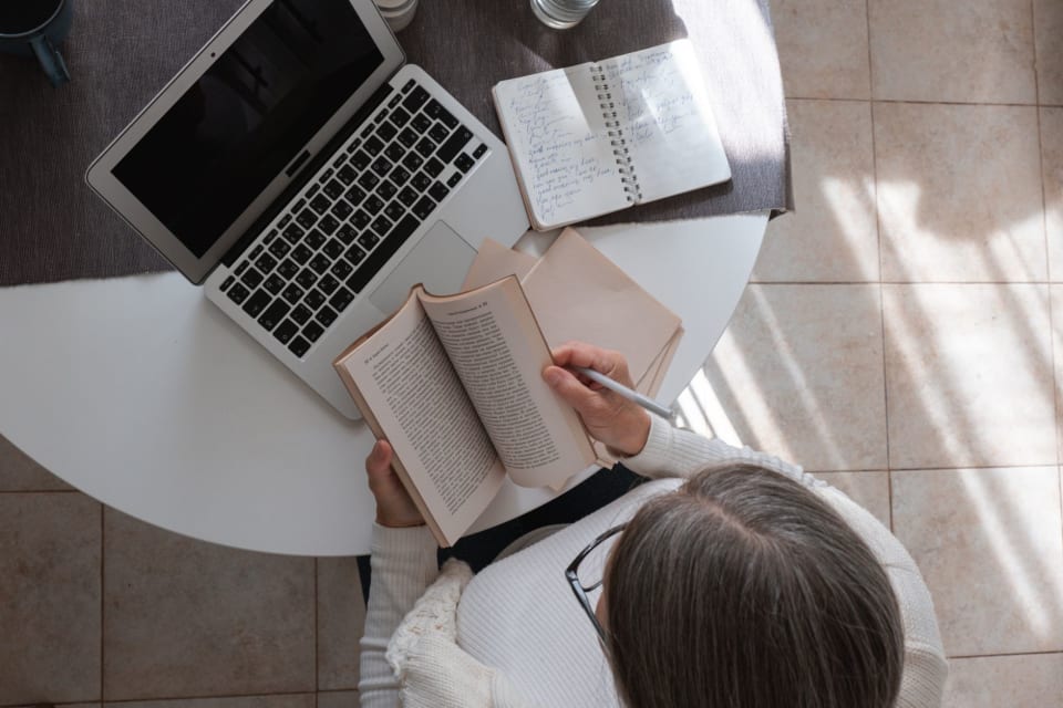 Livro aberto nas mãos de quem lê com uma caneta, vistos de cima, sobre uma mesa com notebook e caderno de anotações