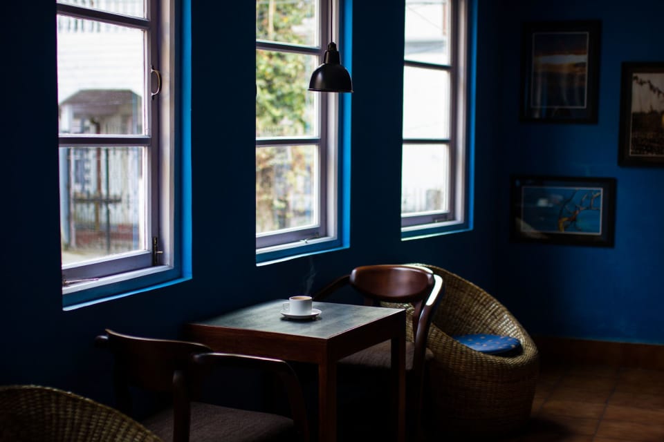 Mesa de madeira com uma xícara junto às janelas de um café de paredes azuis, sob uma luminária.