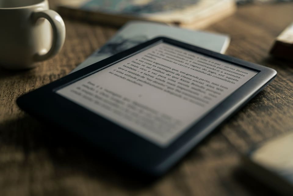 Um leitor de e-book e um livro impresso lado a lado sobre uma mesa de madeira.