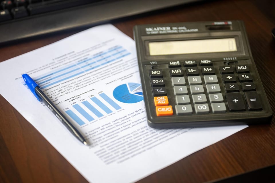 Calculadora, caneta e folhas impressas com colunas de números sobre uma mesa.