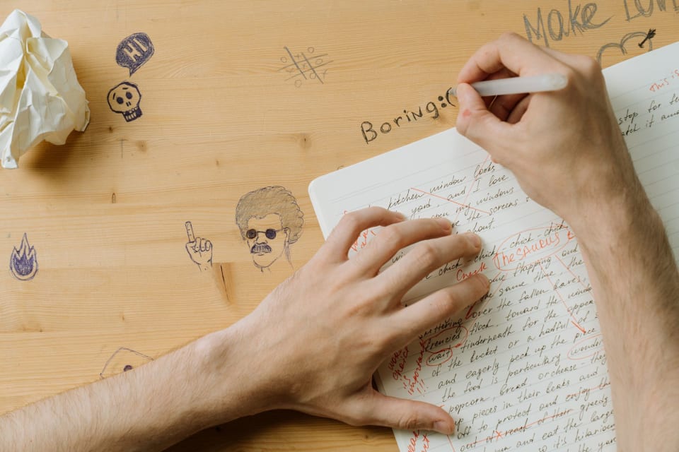 Mãos corrigindo com caneta vermelha um texto escrito à mão num caderno, ao lado de papel amassado e rabiscos na mesa.