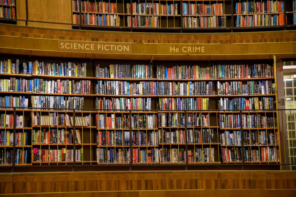 Estantes curvas de biblioteca cheias de livros, com a placa da seção de ficção científica no alto.