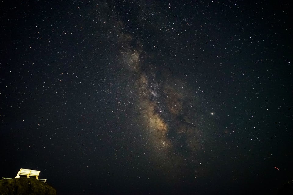 Céu noturno com a faixa da Via Láctea atravessando a imagem e, no canto, a silhueta escura de um morro.