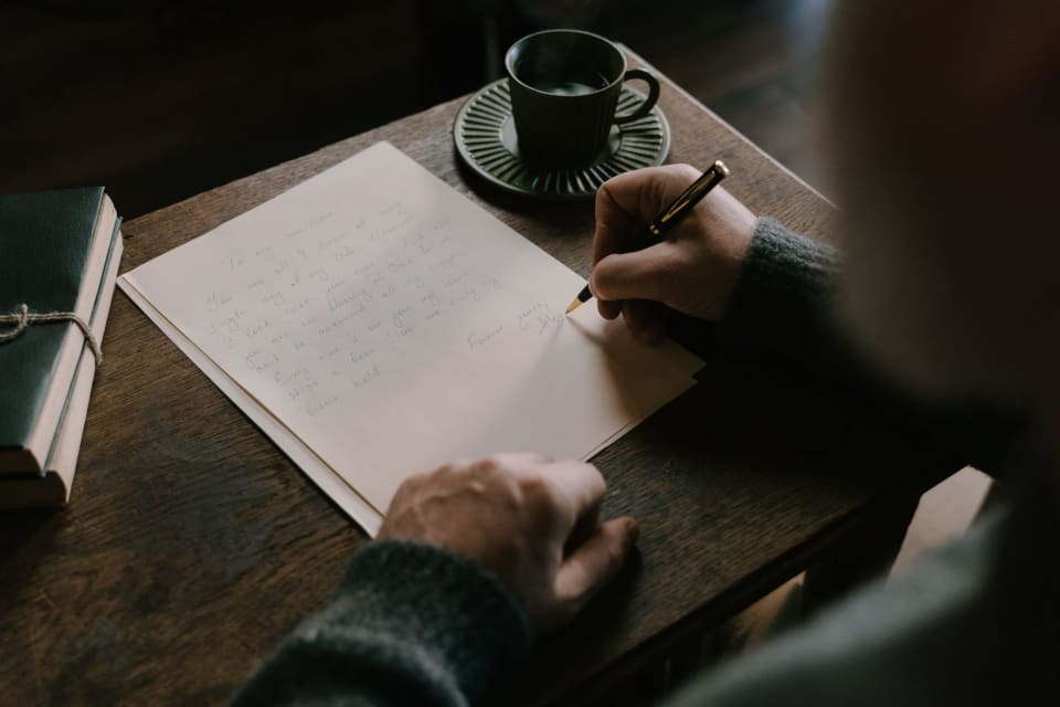Mãos escrevendo à caneta numa folha já meio preenchida, sobre uma mesa de madeira com uma xícara de café e cadernos amarrados com barbante.