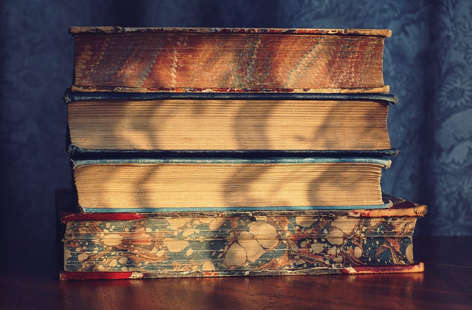 Pilha de livros antigos com cortes marmorizados sobre uma mesa de madeira, sob luz de janela