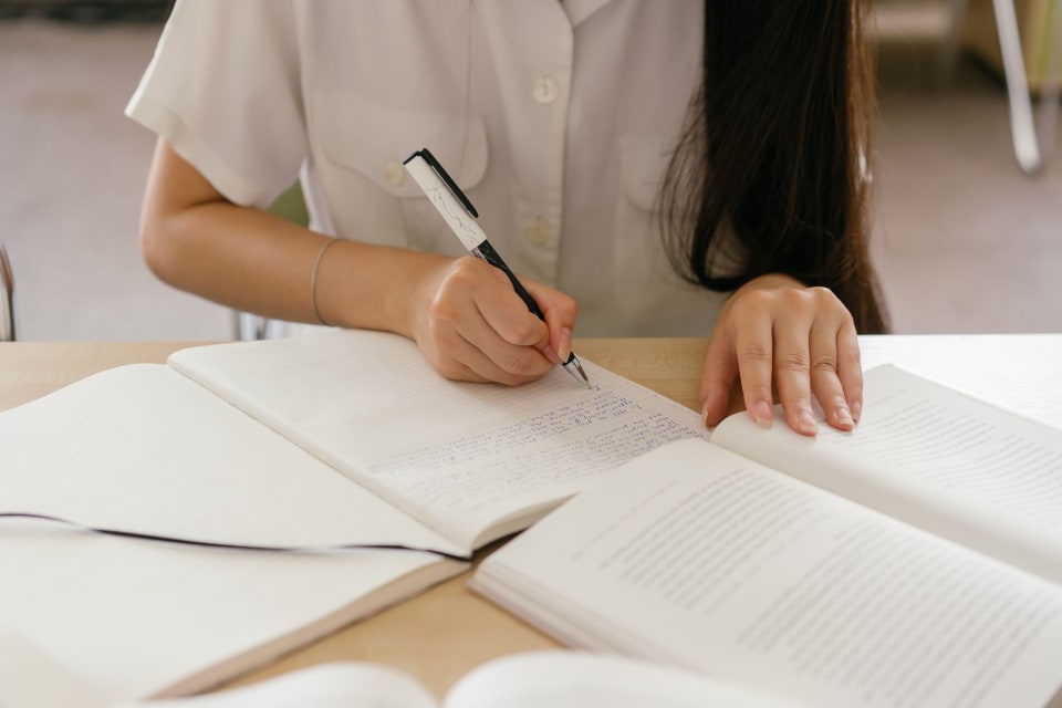 Mão escrevendo anotações num caderno ao lado de um livro aberto, sobre uma mesa