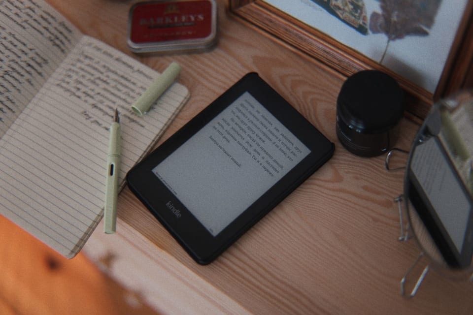 Um leitor de e-book e um livro impresso aberto lado a lado sobre uma mesa de madeira.