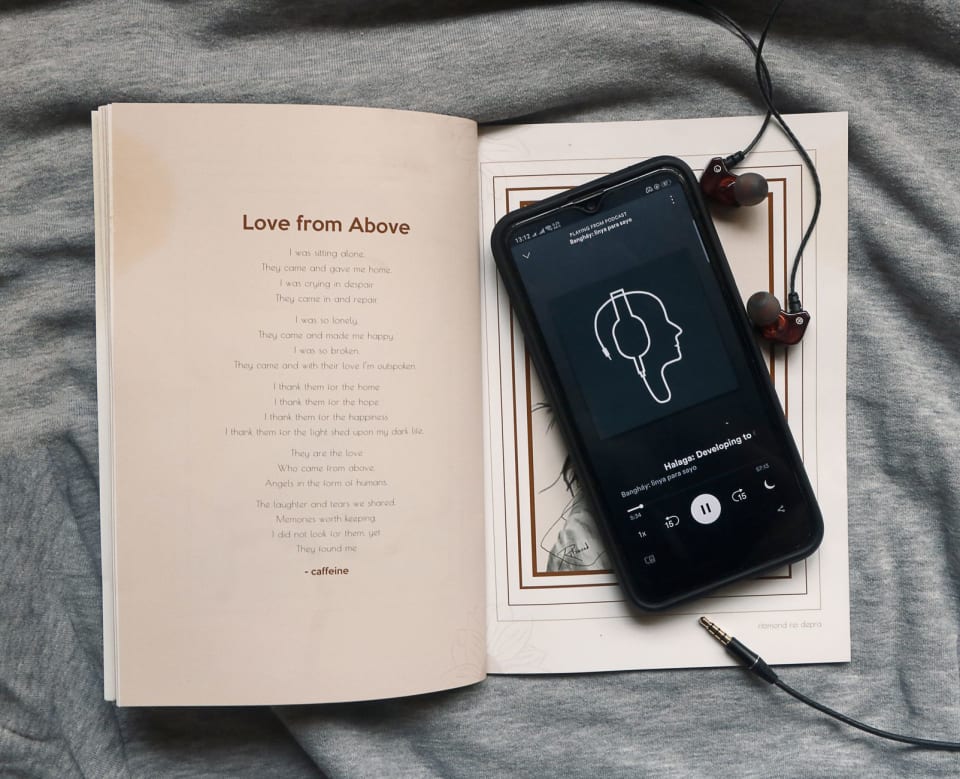 Celular tocando um audiolivro sobre um livro aberto, com fones de ouvido de fio ao lado, num tecido cinza.