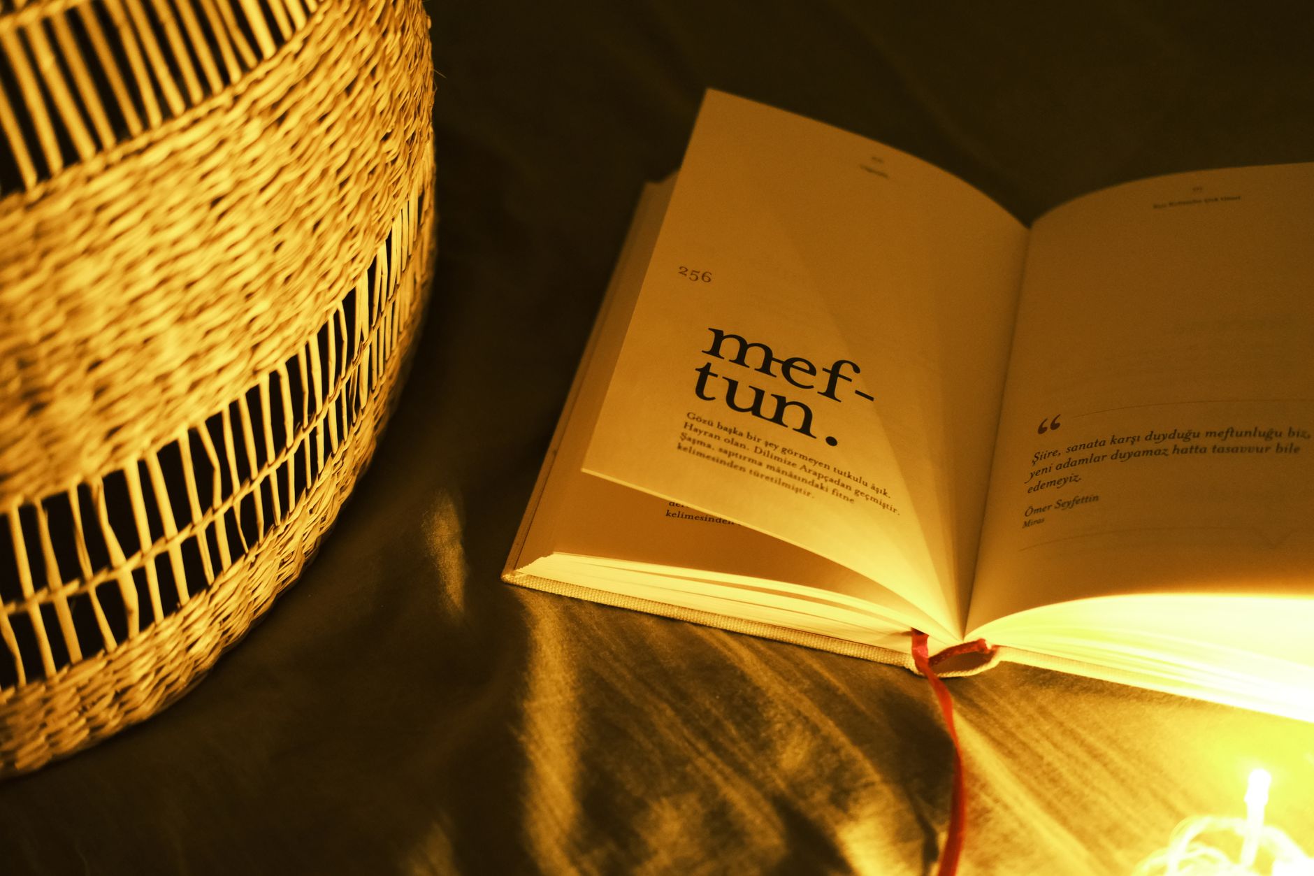 Livro aberto sobre um tecido escuro, na luz quente de uma luminária de palha, com uma página se erguendo e a fita marcadora solta