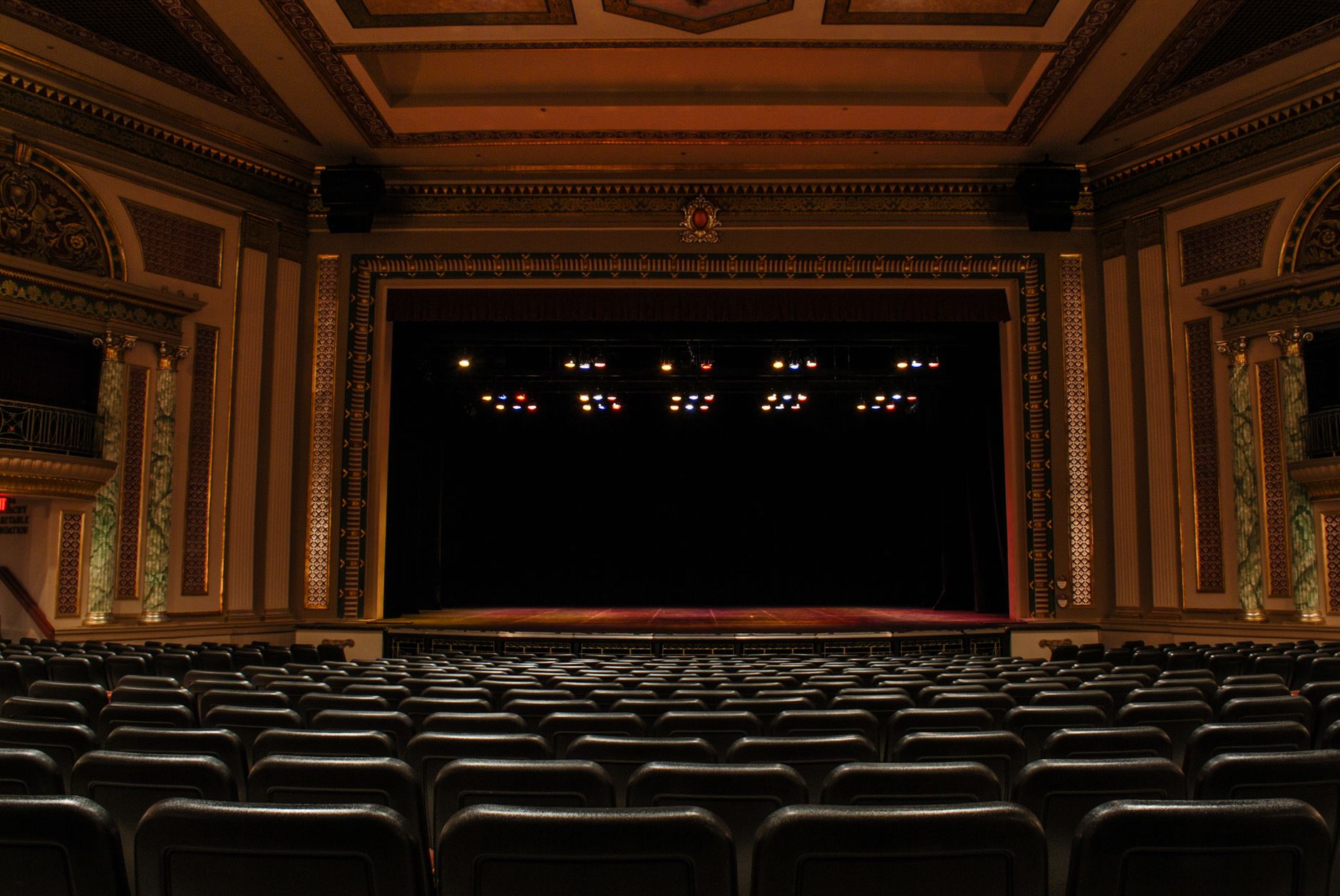 Palco de um teatro antigo, vazio, visto da plateia, com a boca de cena aberta e as luzes de cena acesas no alto