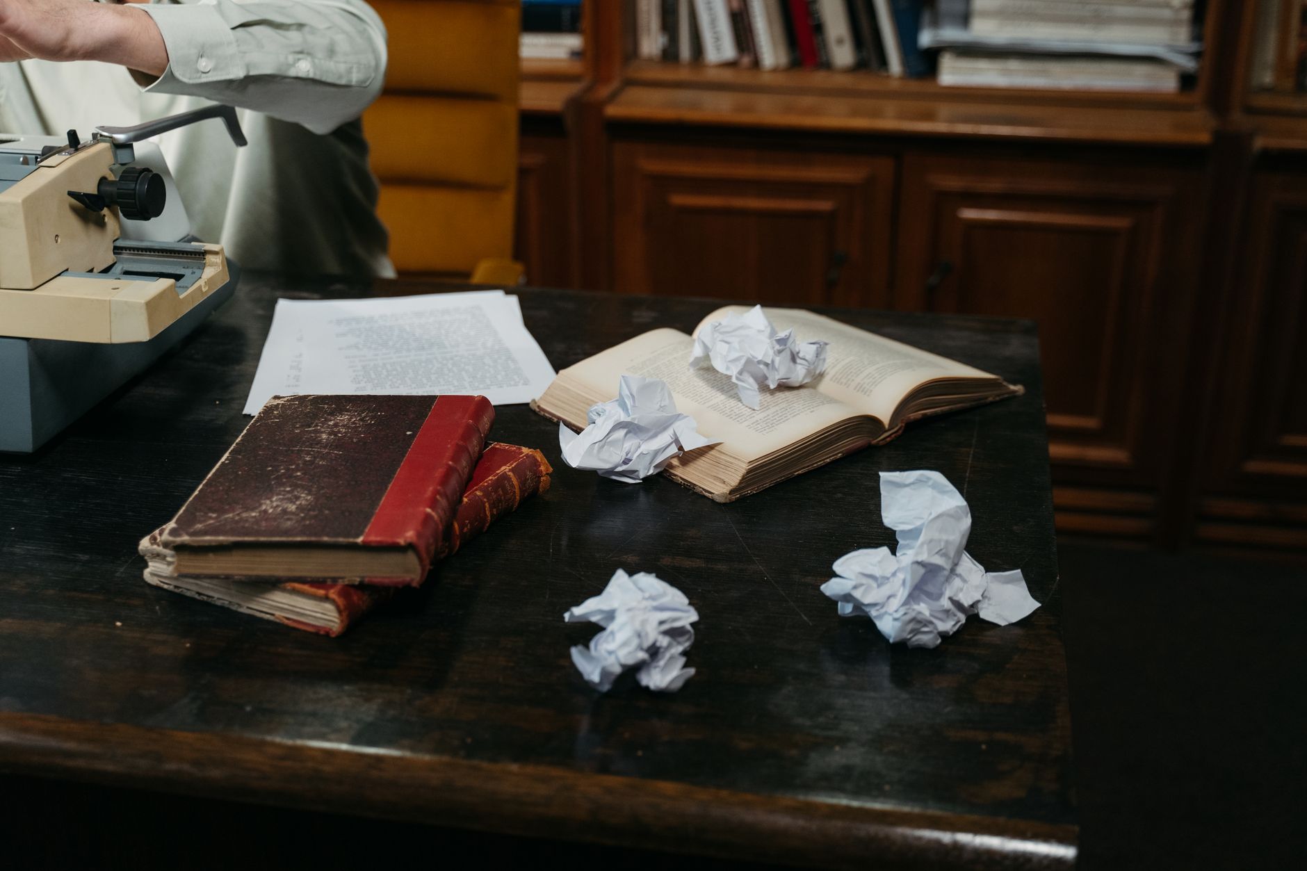 Mesa de madeira escura com uma máquina de escrever, uma folha datilografada, um livro aberto e bolas de papel amassado.
