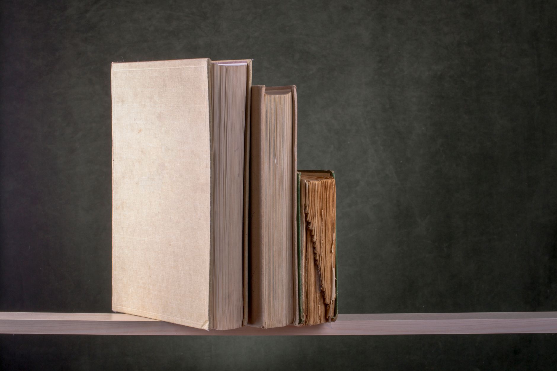 Três livros de pé numa prateleira, do mais grosso ao mais fino, contra uma parede escura.