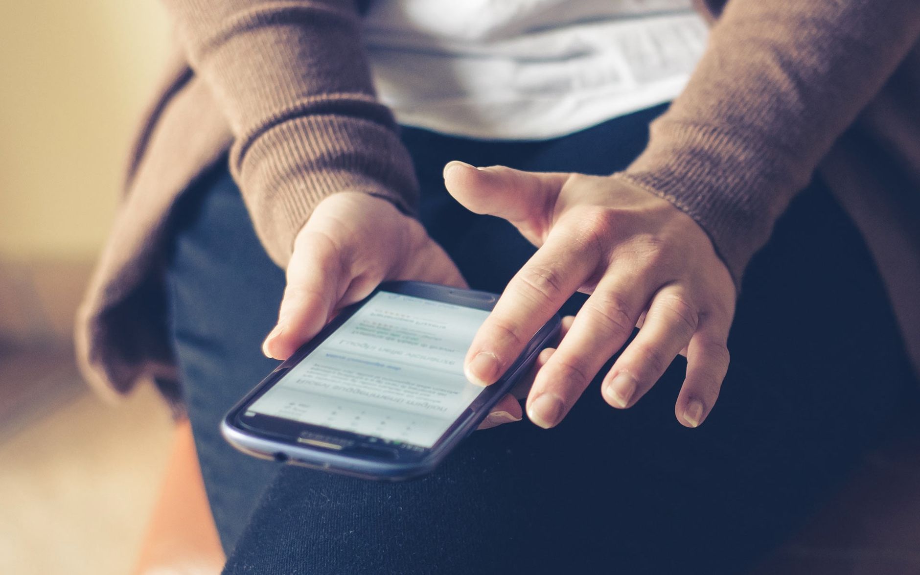 Mãos seguram um celular apoiado no colo, com um texto aberto na tela