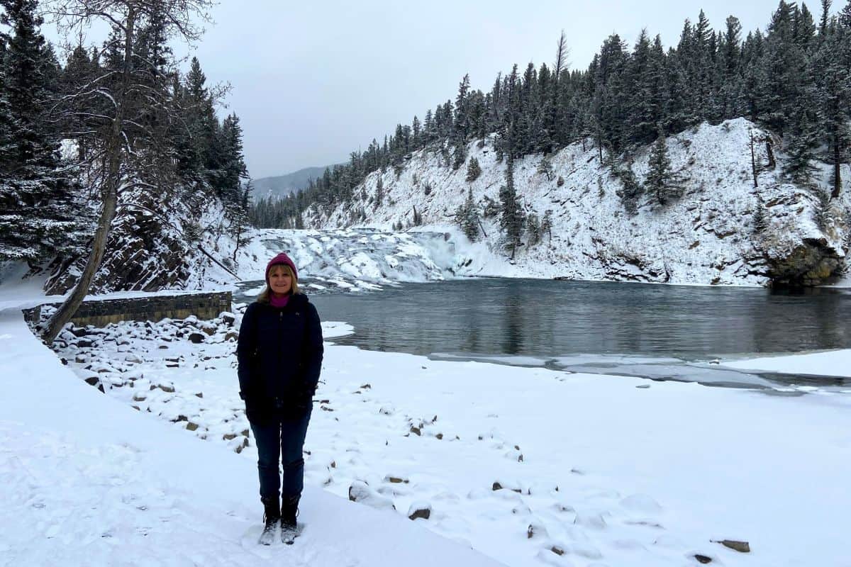 A person on the snowy shore beside open river water at Bow Falls in Banff.