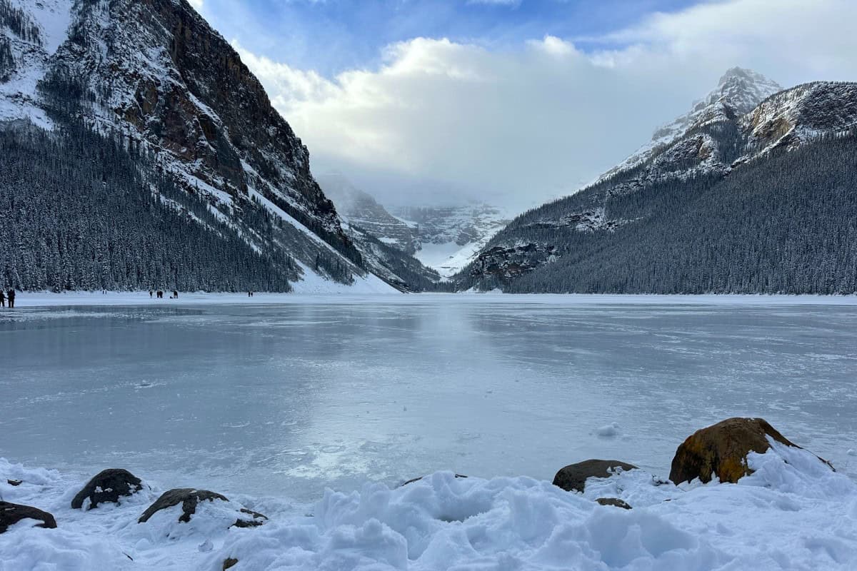 Frozen Lake Louise seen from a snowy, rocky shoreline beneath forested mountain slopes.