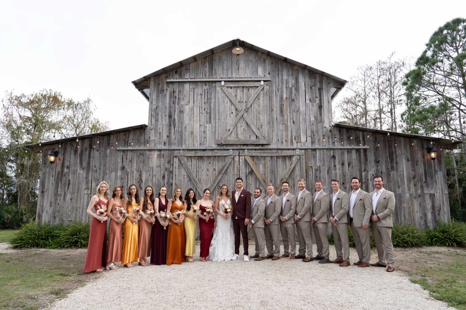 Full 16-person bridal party with bride and groom in front of Misty Morning Barn — bridesmaids in warm autumn tones, groomsmen in tan suits
