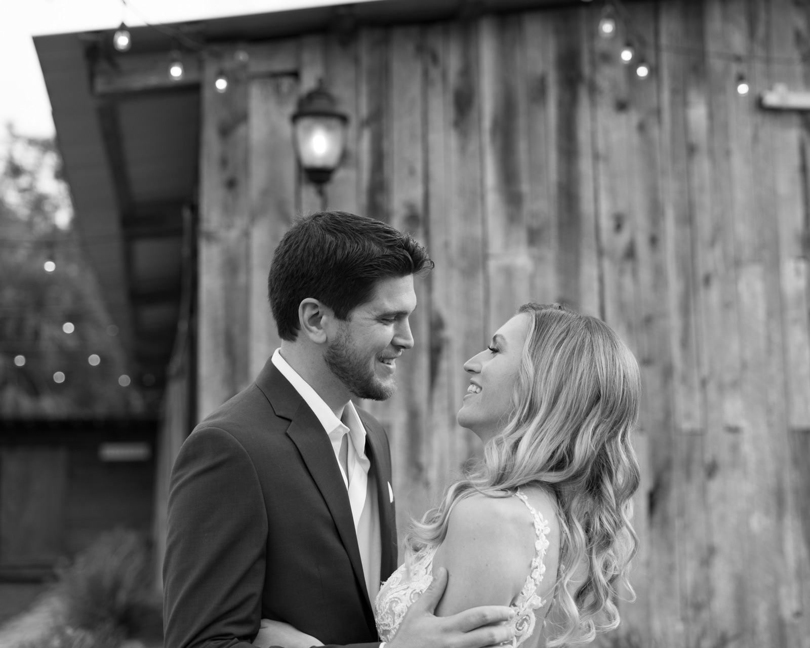 Black and white portrait of Kelli and Bradley laughing together in front of the rustic barn wall with string lights at Misty Morning Barn