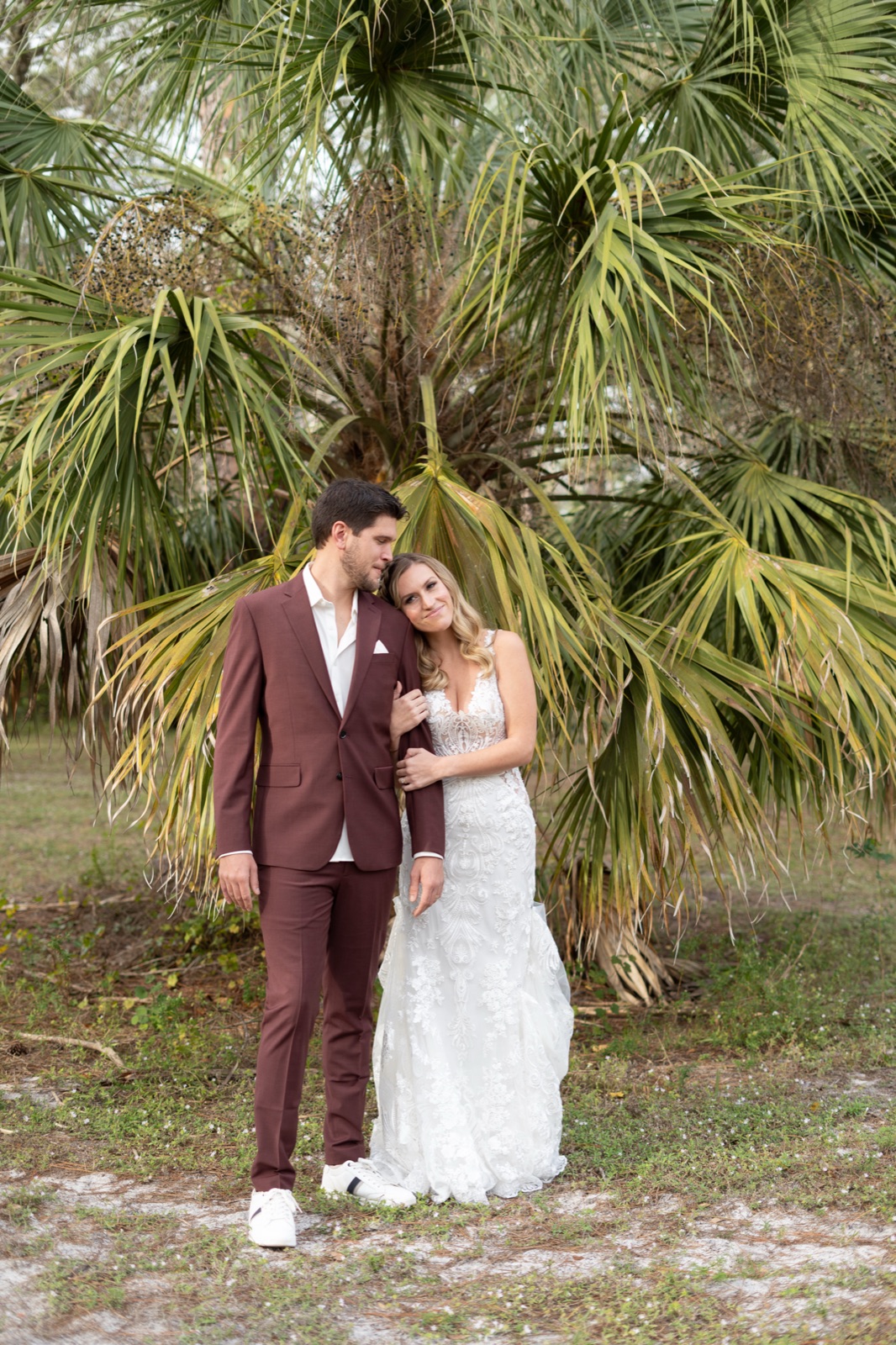 Full-length portrait of Kelli and Bradley under a sabal palm tree at Misty Morning Barn — bride in lace gown, groom in burgundy suit