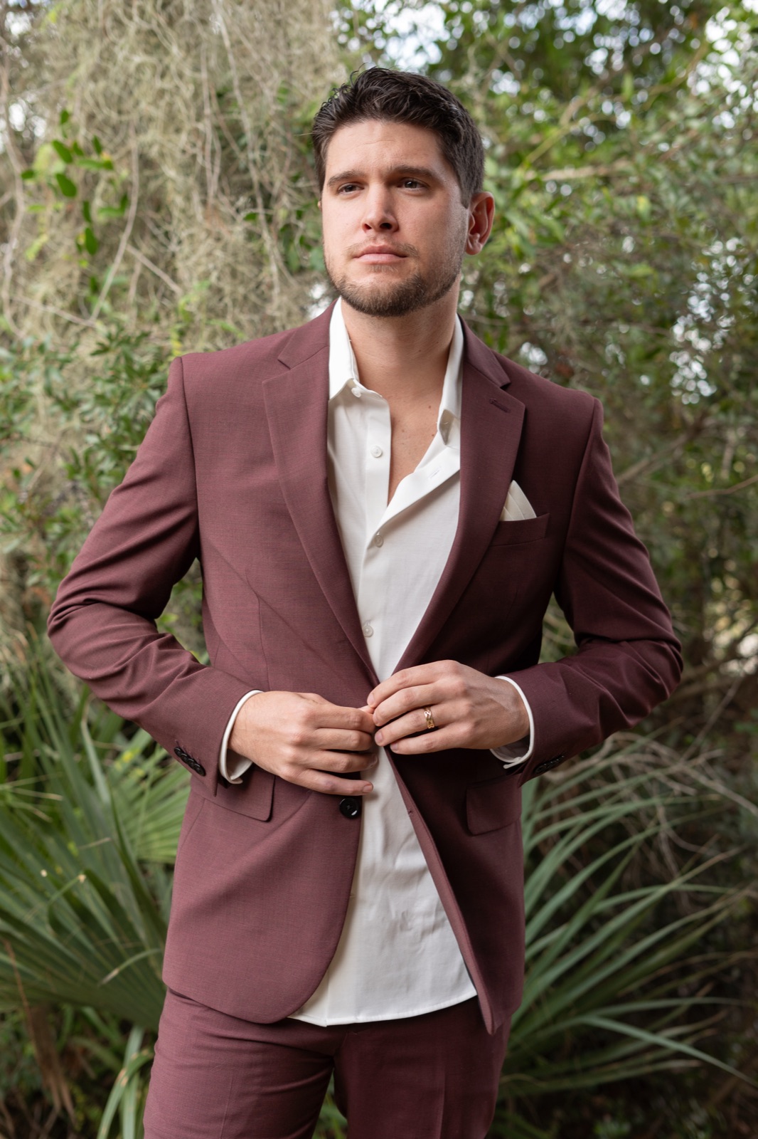 Groom Bradley in burgundy wedding suit with white pocket square buttoning his jacket outdoors at Misty Morning Barn, Fort Myers