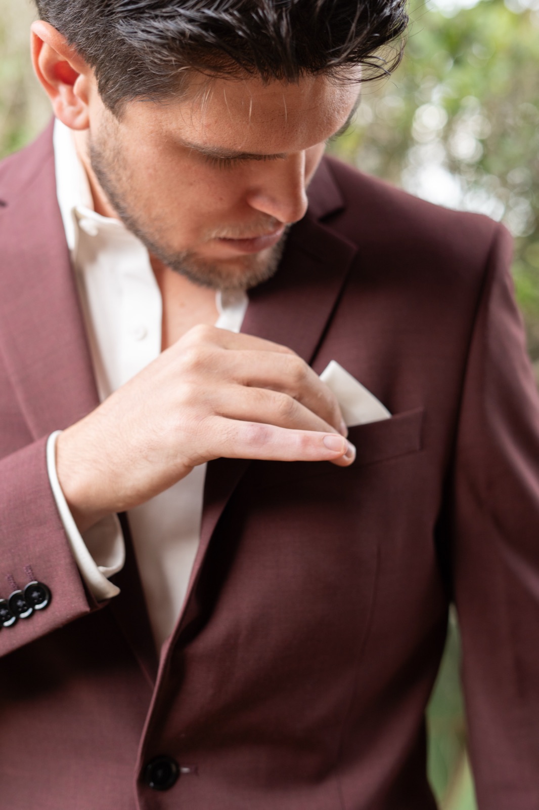 Close-up detail of groom adjusting white pocket square in burgundy wedding suit