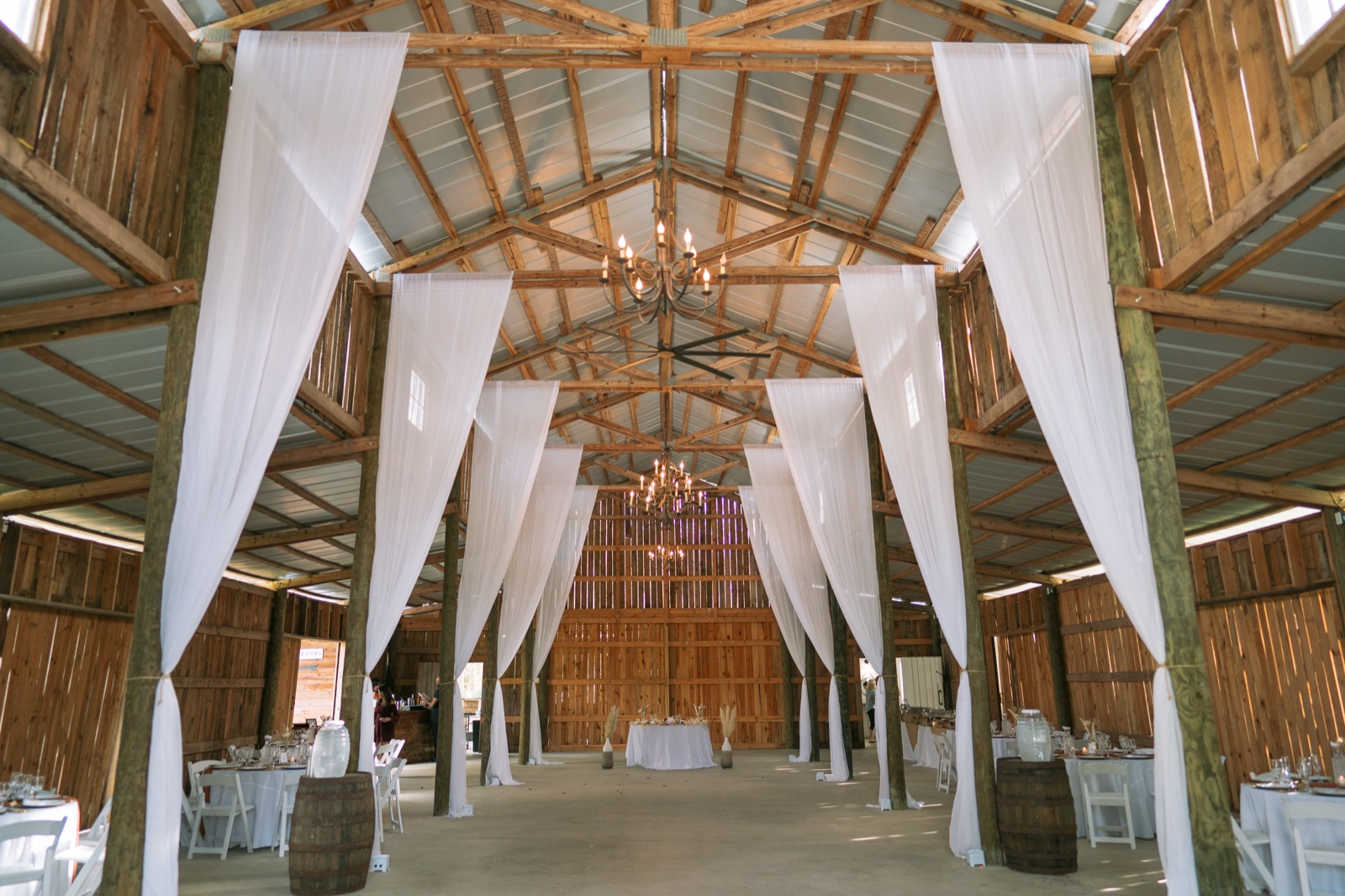 Misty Morning Barn interior with exposed timber beams, white draping, iron chandeliers, and reception tables set for Kelli and Bradley's wedding reception